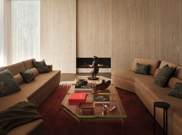 A refined living room with two large tan upholstered sofas facing each other around a glass-topped coffee table. The room features a full-height light timber panelled wall with a centrally inset linear fireplace, sheer curtains and a large window on the left, a deep red rug, dark sculptural side table, and carefully arranged books and decorative objects. The overall atmosphere is warm, sophisticated, and intimate.