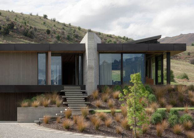 Single-storey contemporary house set into a dry, sloping hillside landscape. The facade combines dark horizontal cladding, large floor-to-ceiling glazed openings, a prominent pale concrete vertical element, and a sharply projecting low roof. A stepped entrance rises through drought-tolerant ornamental grasses and landscaped garden beds.