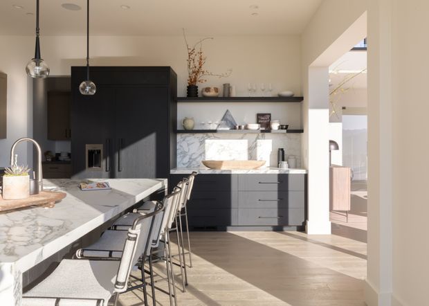 Bright contemporary kitchen with a large veined stone island and integrated seating, dark matte cabinetry, open shelving, marble-look splashback, and pendant lighting. An adjacent room is visible through a wide opening on the right.