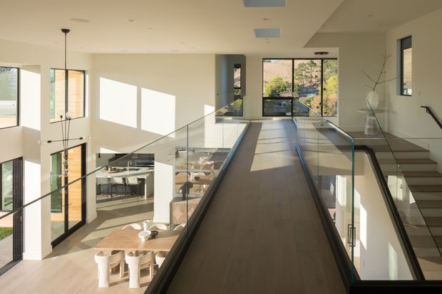 Bright contemporary multi-level interior viewed from an upper landing or bridge walkway. A broad timber-finished circulation bridge with clear glass balustrades overlooks an open-plan dining and kitchen area below. Floor-to-ceiling glazing, high ceilings, a stairway, and strong afternoon sunlight create an airy, spacious atmosphere.