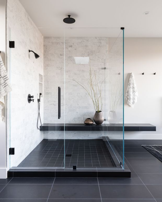 Contemporary bathroom featuring a large frameless glass walk-in shower with black fixtures, a rain showerhead, handheld shower, built-in black bench, and dark tiled flooring. The shower walls are clad in light marble-look tile, with pale painted walls, black accents, and minimalist decorative vessels with branches. A towel rail and bath textiles are visible at the sides.