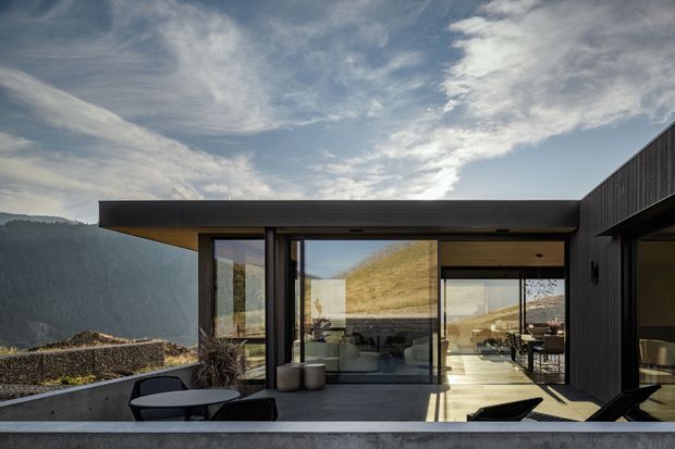 A low-profile contemporary house set against a mountainous landscape, photographed from an exterior terrace. The building has a pronounced flat roof with deep overhangs, dark vertical cladding, extensive floor-to-ceiling glazing, and a concrete terrace edge. Indoor living and dining areas are visible through the glass, while outdoor lounge furniture occupies the foreground.