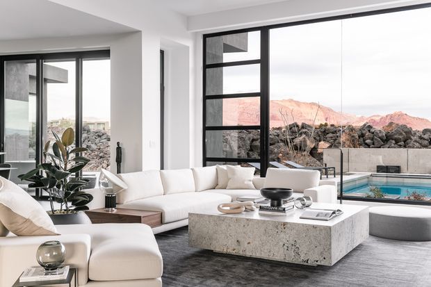 Bright contemporary living room with expansive black-framed floor-to-ceiling glazing overlooking a rocky landscaped setting and pool. The room features white upholstered sofas and lounge chairs, a large textured light-stone coffee table, dark flooring, and a restrained neutral palette. A sculptural indoor plant and minimalist accessories add warmth and visual interest.