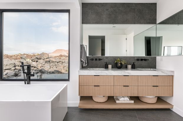 A contemporary bathroom featuring a freestanding white bathtub beside a large black-framed window overlooking a rocky landscape. The room includes a floating light-wood vanity with integrated drawers, a white stone benchtop, dark grey tiled walls and flooring, a full-width mirrored splashback, and black tapware and hardware.
