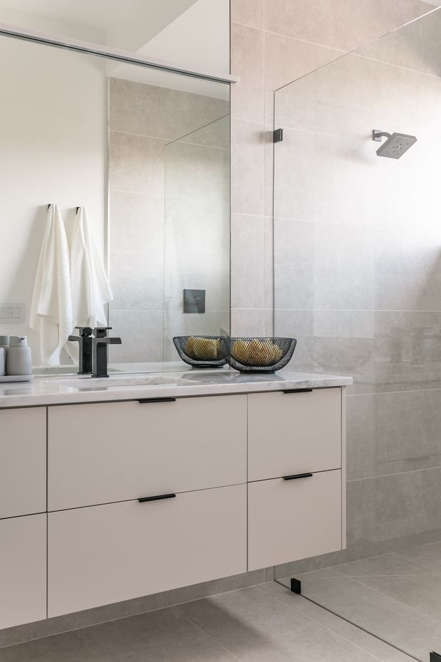 A bright, contemporary bathroom featuring a wall-mounted pale vanity with dark handles, a white stone benchtop, a black geometric mixer tap, and a large frameless mirror. The walk-in shower has large light grey-beige tiled walls and floor, with a square rainfall showerhead and minimal dark hardware. White towels and decorative bowls sit on the vanity.