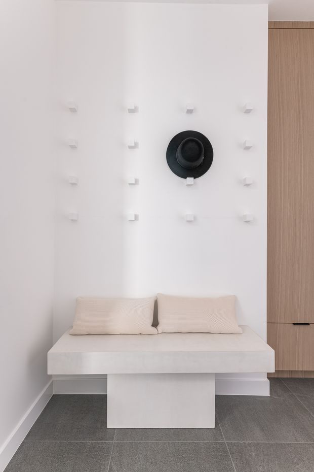 Minimalist bedroom seating/sleeping nook with a low white built-in daybed, two cream pillows, and a black circular wall-mounted accent. The white wall features a regular grid of small square hooks or pegs, with tall light-wood built-in cabinetry at the right. Large grey floor tiles and clean geometric forms create a restrained, contemporary interior.