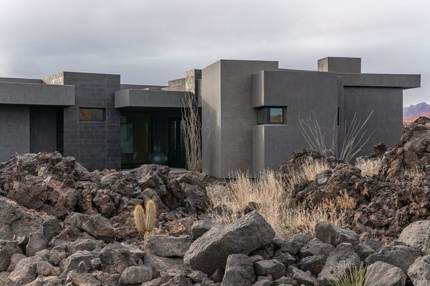 A low, angular dark-grey concrete residence set among rugged volcanic rocks and sparse desert vegetation. The building has a strongly geometric composition of box-like volumes, flat roof planes, narrow horizontal windows, and recessed glazed openings. An overcast sky and rocky terrain create a dramatic, austere landscape setting.