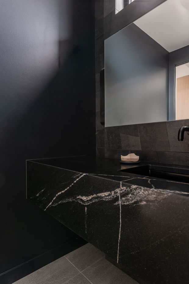 A moody contemporary bathroom with a floating dark stone vanity, integrated black basin and tap, large frameless mirror, and dark tiled wall finish. The space has a dramatic high ceiling with angled architectural geometry and a doorway visible at right.