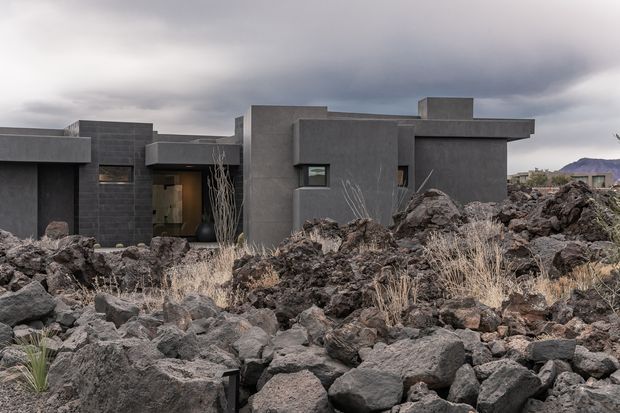 A low, angular contemporary house with dark charcoal concrete or masonry walls, set amid rugged black volcanic rock landscaping. The building features strong horizontal rooflines, projecting box forms, recessed glazing, and a sheltered entry. Dry grasses and sparse drought-tolerant planting sit among the lava rocks beneath an overcast sky.