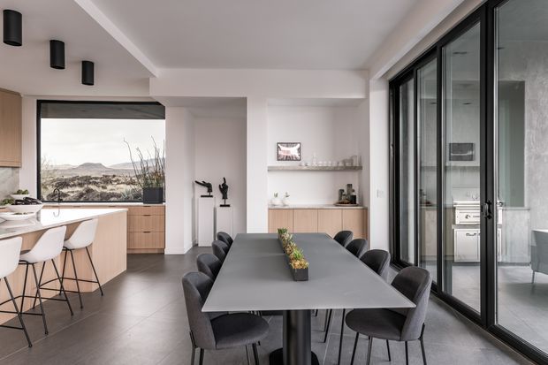 A bright, contemporary open-plan kitchen and dining area with a long dark dining table surrounded by upholstered chairs. The kitchen features pale timber cabinetry, a light island benchtop, integrated appliances, black fixtures and fittings, and large black-framed glazed doors along one side. White walls and ceilings, dark flooring, and minimalist open shelving create a clean, refined interior. A dry, rocky landscape is visible through the window.
