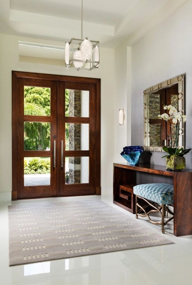 Bright, refined residential entry foyer with dark timber double doors featuring inset glazing, a built-in timber console and bench beneath a large decorative mirror, a patterned upholstered stool, contemporary pendant light, and a pale patterned area rug over glossy light flooring. Garden greenery is visible through the entry glazing.