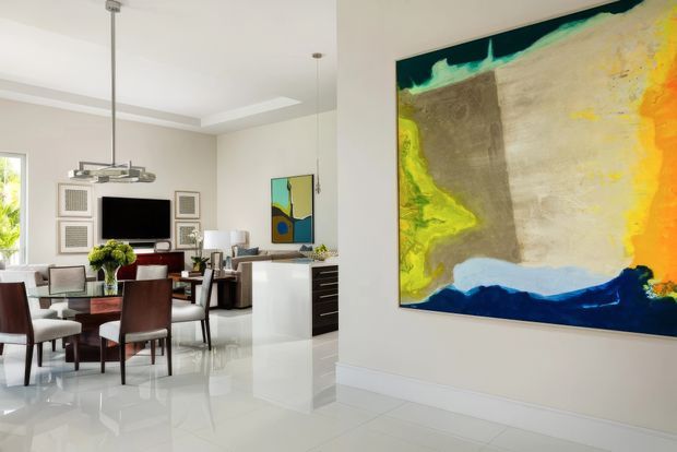 Bright open-plan contemporary interior featuring a dining area, living room, and kitchen zone. The room has glossy white flooring, pale walls, white cabinetry, dark wood dining furniture, recessed ceiling detailing, and a large colourful abstract artwork as a focal point.