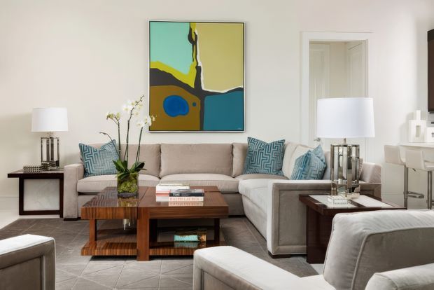 A refined contemporary living room with a large L-shaped upholstered sectional sofa, matching armchairs, patterned blue accent cushions, and a dark timber coffee table. White walls and trim create a bright neutral backdrop, complemented by paired table lamps, a sculptural orchid arrangement, and a bold abstract artwork above the sofa. An open doorway leads to an adjacent space.