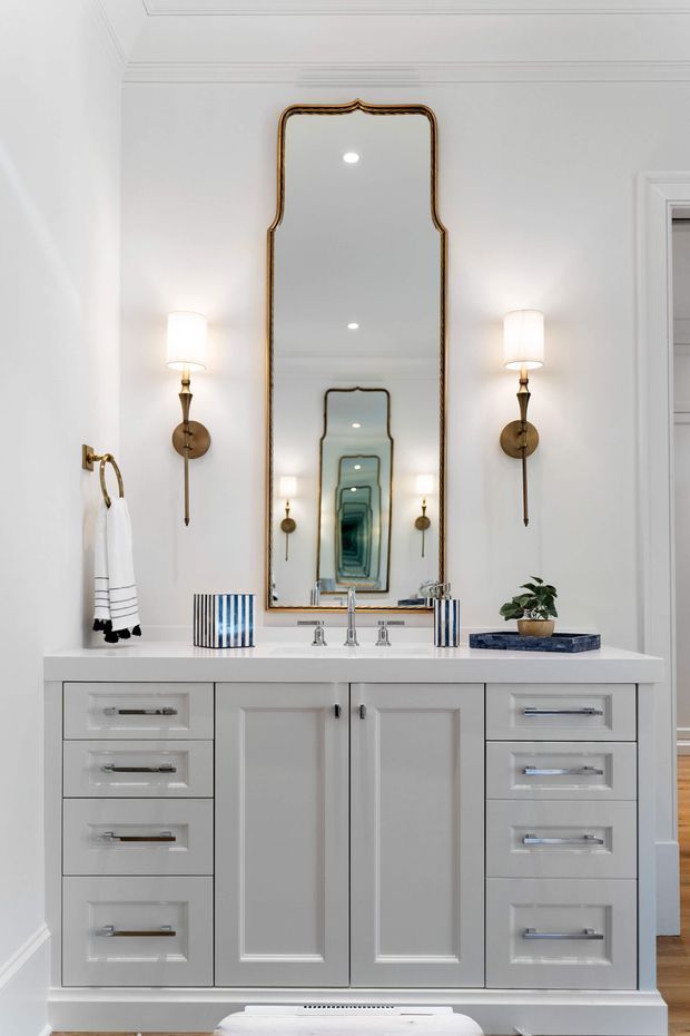 Bright, refined bathroom vanity featuring a large ornate gold-framed mirror, white painted panelled cabinetry with chrome hardware, a white benchtop, twin wall sconces, and a central basin with polished chrome tapware. The mirror creates a strong repeating reflection effect, while subtle wall mouldings and timber flooring add traditional architectural detail.