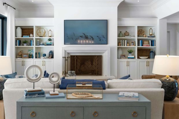 A refined living room viewed from behind a large upholstered sofa, centred on a white-trimmed fireplace with a wall-mounted television above. Built-in white shelving and cabinetry flank the fireplace, displaying books, ceramics and accessories. A blue-grey console with brass-toned drawer pulls occupies the foreground, while layered blue, white and neutral furnishings create a polished, comfortable interior.