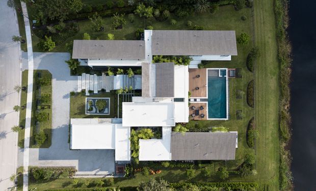 Aerial overhead view of a large contemporary residential property arranged around landscaped courtyards and a rectangular swimming pool. The home comprises several interconnected low-pitched, flat-roofed and tiled-roof volumes, with pale exterior surfaces, extensive lawns, mature planting, paved paths, and a driveway along the street edge. A dark waterway or pond borders the property on the right.