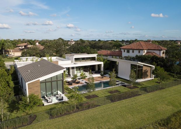 Aerial view of a large contemporary residential property with several connected flat-roof and low-pitched structures arranged around a landscaped courtyard and swimming pool. The home features white rendered walls, extensive floor-to-ceiling glazing, a dark ribbed roof on one pavilion, timber-toned wall panels, paved terraces, lawn, and perimeter fencing. Traditional tiled-roof neighbouring homes and dense greenery surround the site.