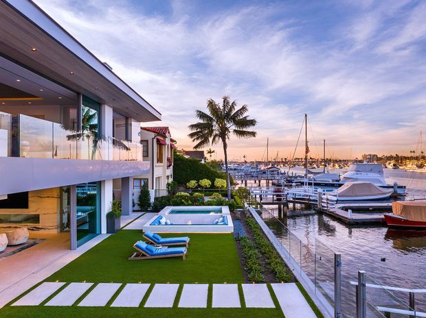 Waterfront exterior view of a contemporary multi-level residence with broad floor-to-ceiling glazing, projecting upper-level balcony, timber soffit, manicured artificial-grass terrace, rectangular swimming pool, stepping-stone path, and direct outlook over a marina with yachts and docks.