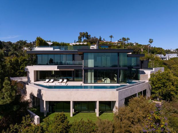 Aerial exterior view of a large contemporary multi-level residence set among mature landscaping on a sloped site. The home features broad floor-to-ceiling glazing, dark framed windows, pale rendered or concrete-looking walls, projecting roof planes, extensive terraces, and a swimming pool along the upper outdoor level. Palm trees and neighbouring homes are visible in the background.