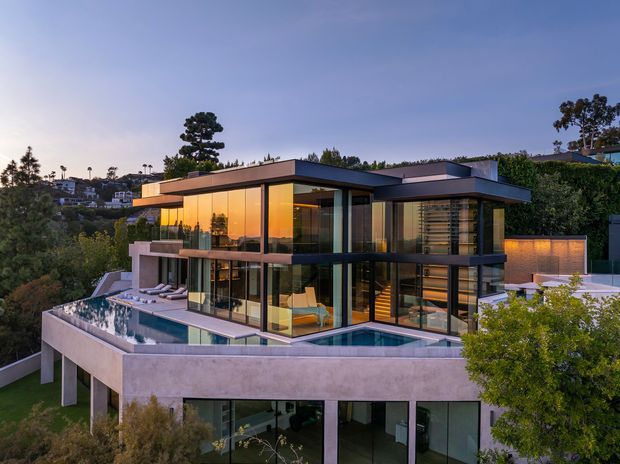 A contemporary multi-level residential house on a sloping, elevated site, photographed at dusk. The home features extensive floor-to-ceiling glazing, projecting flat roof planes, dark window frames, warm interior lighting, concrete terraces and retaining walls, and a swimming pool wrapping around the upper-level outdoor terrace.