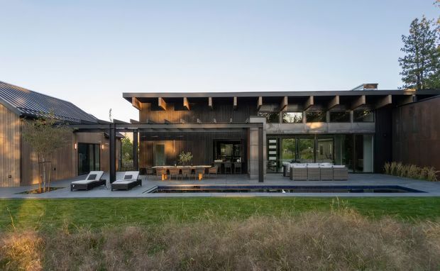 A contemporary single-storey residence viewed from the landscaped rear garden, with a dark timber-clad facade, broad low-pitched roof, deep exposed timber eaves, extensive glazing, a covered outdoor dining and lounge area, and a long narrow reflecting or swimming pool along the terrace.