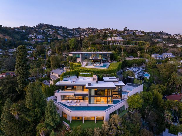 Aerial view of a large contemporary multi-level residence set into a lush, densely vegetated hillside suburb. The home features flat roof planes, extensive floor-to-ceiling glazing, angular terraces, pale rendered walls, landscaped gardens, and a prominent swimming pool. Additional modern homes are visible across the surrounding hillside.