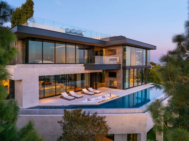 A multi-level modern residence set into a landscaped site, featuring broad floor-to-ceiling glazing, projecting flat roof planes, pale rendered or concrete walls, dark window frames, and a rooftop terrace with glass balustrades. A swimming pool occupies the foreground terrace, with outdoor loungers and built-in seating visible around it.