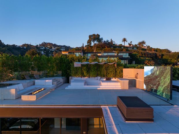 A sophisticated rooftop terrace overlooking a leafy hillside residential setting. The expansive paved roof deck features built-in white lounge seating, a linear fire feature, a large outdoor screen, sculptural shade canopies, and a dark wood-toned low table or storage element. Glass doors and glazing below connect the terrace to the building interior, while warm evening light creates a refined entertainment atmosphere.