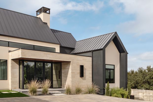 Exterior architectural photograph of a contemporary multi-wing house with steep gabled and sloped standing-seam metal roofs, pale stone-clad walls, dark contrasting wall panels, and extensive black-framed glazing. A covered stone entry or outdoor living area opens onto a paved terrace with ornamental grasses and landscaped planting.