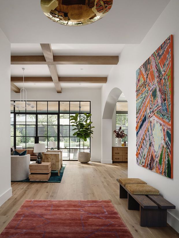 A refined open-plan living room with pale timber flooring, white walls, exposed timber ceiling beams, and a large black-framed glazed wall overlooking greenery. The space includes a curved cream sofa, low timber coffee tables, a central potted tree, built-in timber cabinetry, a long dark timber bench with woven seats, and a large colourful abstract artwork. A rounded metallic ceiling light is partly visible overhead, adding a sculptural accent.