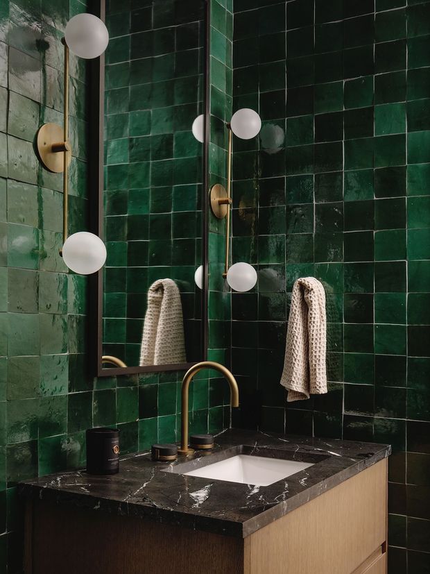 A compact, richly coloured bathroom vanity with deep green glossy square wall tiles, a dark veined stone benchtop, and warm wood-grain cabinetry. A brass-toned wall-mounted basin mixer and matching hardware sit beneath a dark-framed mirror, flanked by spherical wall lights. A textured neutral towel hangs beside the mirror. The space has a moody, refined, slightly vintage-inspired interior aesthetic.