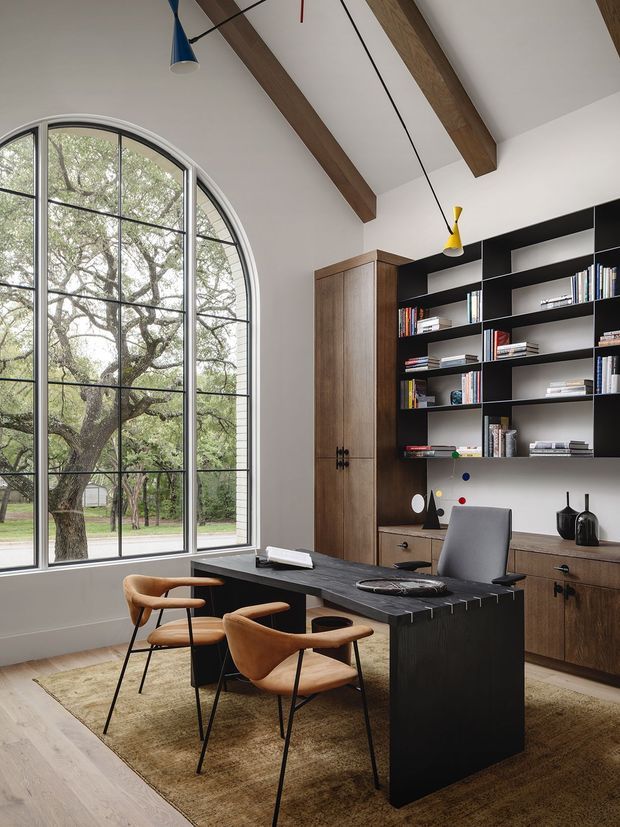 A refined home office with a dramatic double-height feel, featuring a large arched steel-framed window, exposed timber ceiling beams, a black desk with meeting chairs, a grey office chair, and extensive dark built-in shelving and timber cabinetry. Warm wood flooring and a textured neutral rug balance the monochromatic joinery, while colourful pendant lights add small accents.