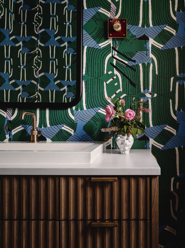 Close-up of a richly patterned powder-room vanity. A white stone countertop with an integrated rectangular basin and brushed brass tap sits above dark vertically fluted timber cabinetry with brass pulls. The wall is covered in a bold graphic botanical-style wallpaper in deep green, blue, cream and black, with a black-framed mirror, wall-mounted decorative object and floral arrangement adding detail.