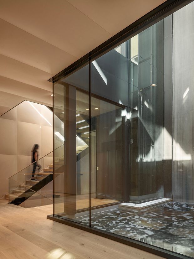 A refined contemporary interior atrium with a glazed internal courtyard or lightwell. Floor-to-ceiling glass partitions frame a dark textured vertical wall and shallow reflective water feature, while a timber staircase with slim metal balustrades rises beside pale timber flooring. Warm recessed lighting and strong natural sunlight create layered reflections and shadow patterns.