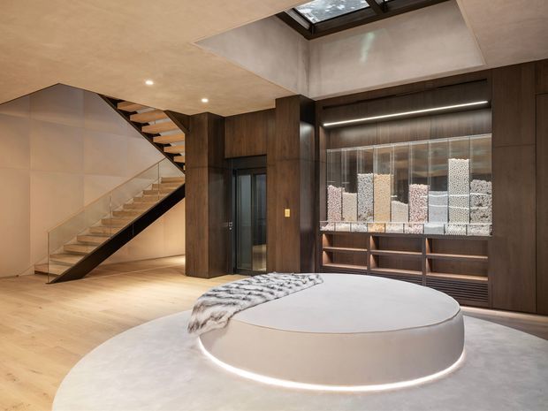 A sophisticated contemporary interior featuring a sculptural circular raised platform or seating island in the foreground, a timber staircase with open risers, dark timber wall panelling and built-in cabinetry, a concealed lift or doorway, and a large glazed display feature. A skylight above the stair and recessed linear lighting create a warm, refined atmosphere. The space appears to function as an entry or circulation area with a dramatic architectural focal point.