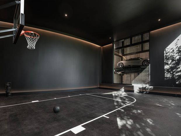 A dark, dramatic indoor basketball court or sports garage with matte charcoal walls and ceiling, a basketball hoop mounted at the left, white court markings on a dark floor, and a large glazed opening revealing a parked sports car on a raised platform beyond. Warm concealed perimeter lighting and dappled daylight create strong contrast.