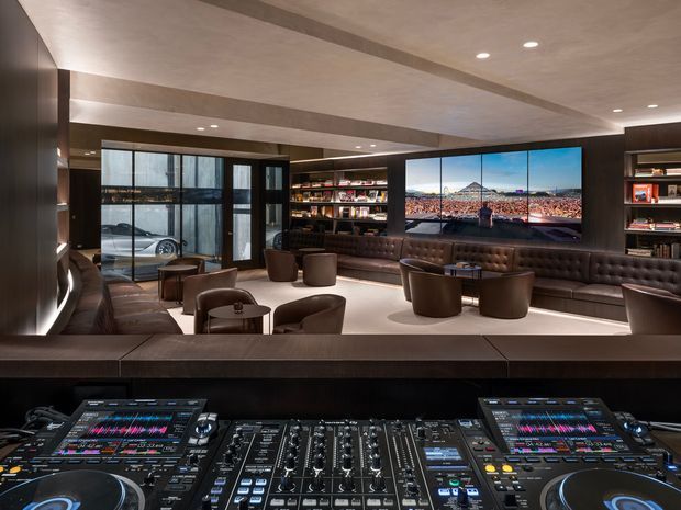 A dark, sophisticated entertainment lounge or DJ booth overlooking a spacious seating area. The foreground is dominated by professional DJ equipment and a mixing console, while the background features leather banquette seating, lounge chairs, extensive dark timber shelving, a large wall-mounted display, recessed lighting, and glazed doors with a vehicle visible beyond. The interior has a refined, moody hospitality or club-like atmosphere.