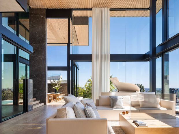 A luxurious double-height living room with floor-to-ceiling glazing, expansive ocean views, pale upholstered sofas, a sculptural white lounge chair, a large light-wood coffee table, and a dramatic fluted vertical feature column. Dark marble-look wall panels and black window framing contrast with the warm timber slatted ceiling and timber flooring. The space is bright, airy, and highly refined.