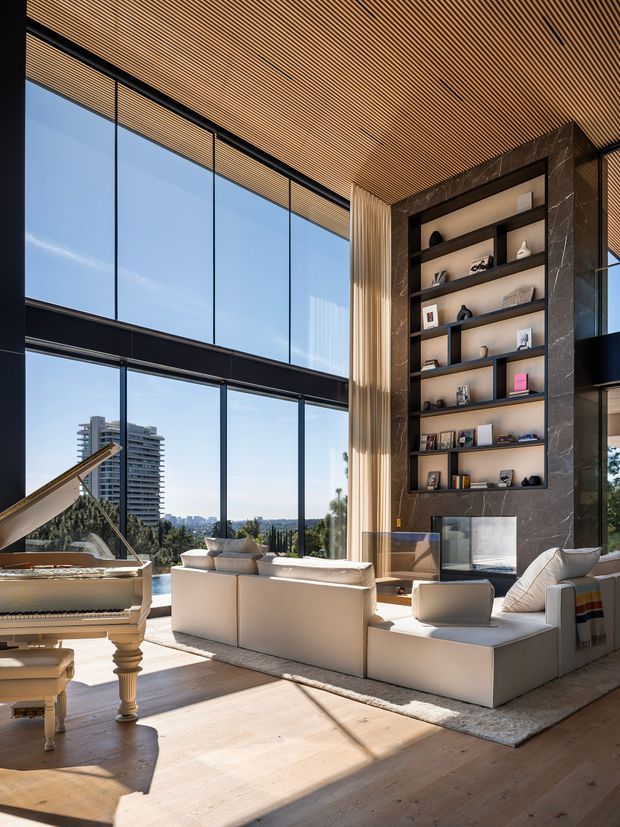 A bright, luxurious double-height living room with expansive floor-to-ceiling glazing, a warm slatted timber ceiling, pale timber flooring, and a large contemporary white sectional sofa arranged around a fireplace. A dramatic dark stone built-in shelving wall displays decorative objects and books, while a grand piano occupies the foreground. The glazing overlooks a leafy urban landscape, and strong sunlight creates a warm, airy atmosphere.
