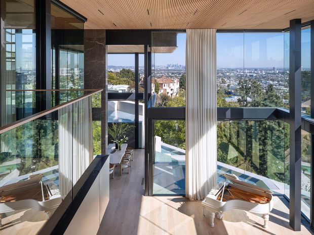 A bright, elevated interior overlooking a dense cityscape, featuring extensive floor-to-ceiling glazing, slim dark window frames, a pale timber slatted ceiling, light timber flooring, full-height sheer curtains, and a white grand piano. A glass balustrade and open stair or landing connect to the lower level, with trees and rooftops visible beyond.