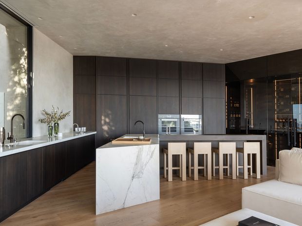 Contemporary open-plan kitchen and entertaining space with dark wood floor-to-ceiling cabinetry, a white marble-look island, integrated breakfast bar seating, and a glazed wine display cabinet. The room has a warm timber floor, textured plaster ceiling, expansive glazing, and a neutral upholstered sofa in the foreground.