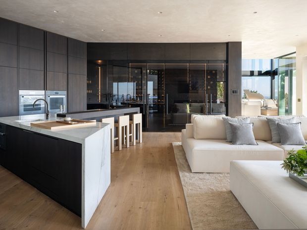 Open-plan contemporary kitchen and living area with dark full-height cabinetry, integrated appliances, a large pale stone island, timber flooring, and a white sectional sofa. A glass-fronted dark bar or display area sits behind the kitchen, while bright floor-to-ceiling glazing opens the living space to an exterior view.