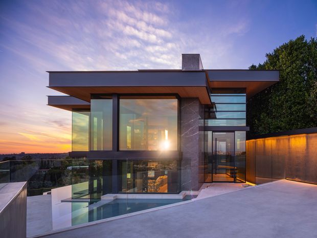 A sophisticated multi-level modern house photographed at sunset, featuring broad cantilevered flat roof planes, extensive floor-to-ceiling glazing, dark metal-framed windows, a tall stone-clad vertical wall, and a rooftop terrace with glass balustrades. A partially visible lower-level swimming pool and illuminated interior spaces reinforce the indoor-outdoor connection. The setting appears elevated, with distant landscape views beyond the property.