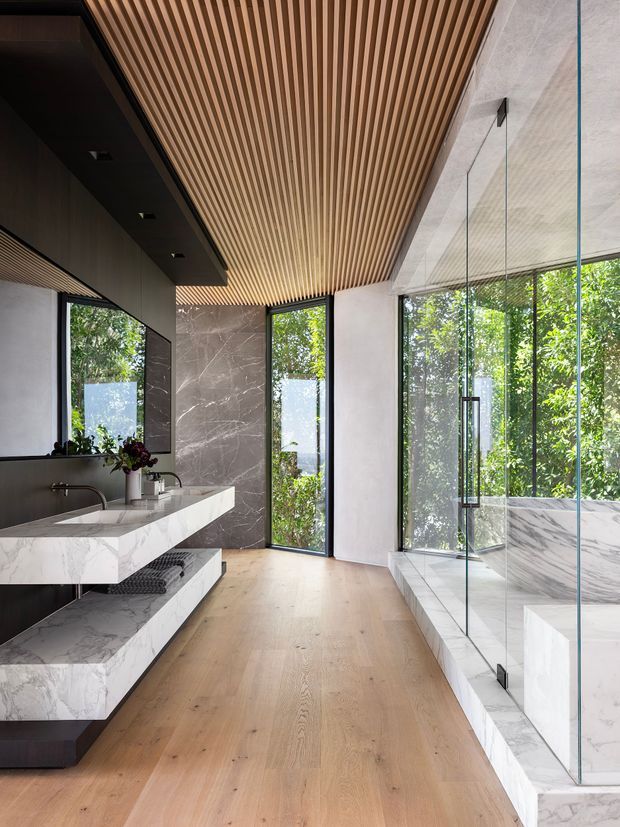 Contemporary luxury bathroom with a floating double vanity in veined white marble, dark wall cabinetry, and a spacious glass-enclosed shower or wet area. The room features a warm linear timber-slatted ceiling, light timber flooring, large black-framed glazed openings, and abundant greenery visible outside. Clean architectural lines and natural light create a refined, bright, indoor-outdoor atmosphere.