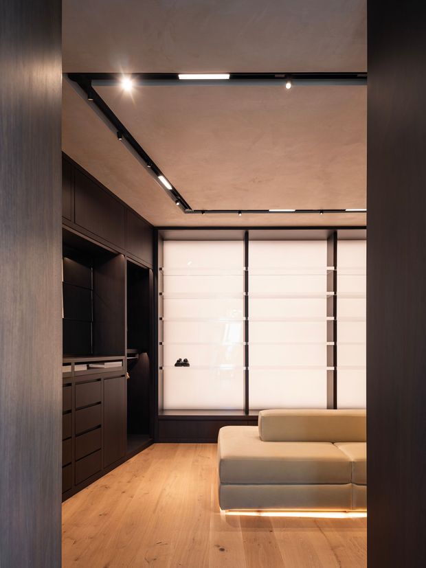 A compact, refined interior viewed through a dark timber-framed opening. The room features full-height dark built-in cabinetry and drawers along the left wall, warm timber flooring, a large translucent segmented garage-style door or glazed wall at the rear, and a beige upholstered sofa with subtle underlighting. Black recessed track lighting runs across the warm plaster ceiling, creating a contemporary, softly illuminated space.