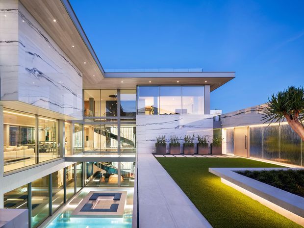 Twilight exterior view of a luxurious multi-level contemporary residence organized around a central glazed courtyard and visible swimming pool. The building features expansive floor-to-ceiling glazing, a flat roof with broad cantilevered eaves, dramatic white veined stone-clad walls, warm timber soffits, integrated lighting, and manicured landscaping with a lawn and raised planter.
