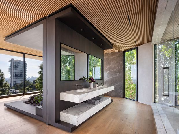 A luxurious contemporary open-plan bathroom with a double floating vanity finished in veined white stone, dark timber or charcoal wall cabinetry, and a large marble-look feature wall. The room has warm timber flooring and a slatted timber ceiling, extensive floor-to-ceiling glazing, and a glass-enclosed area on the right. A leafy outdoor view and a high-rise building are visible through the windows.