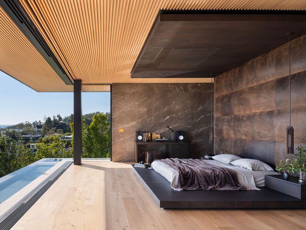 A luxurious contemporary bedroom opening onto a spacious terrace, with a low platform bed, dark bedding, integrated bedside surfaces, and a compact media console against a richly veined dark stone wall. The room features warm timber flooring and a striking slatted timber ceiling, dark metal posts and trim, extensive open glazing, and a broad outdoor view. The palette is warm wood, dark brown, charcoal, and neutral bedding, creating a refined indoor-outdoor atmosphere.