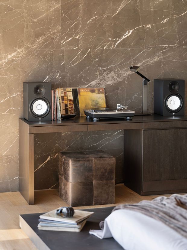 A refined bedroom corner featuring a dark wood console with integrated drawers and cabinetry, a turntable, vinyl records, and two speakers. The console sits against a dramatic brown-grey veined stone wall, with a leather ottoman beneath. Part of the bed and a dark coffee table with books and headphones are visible in the foreground, creating a warm, sophisticated music-focused retreat.