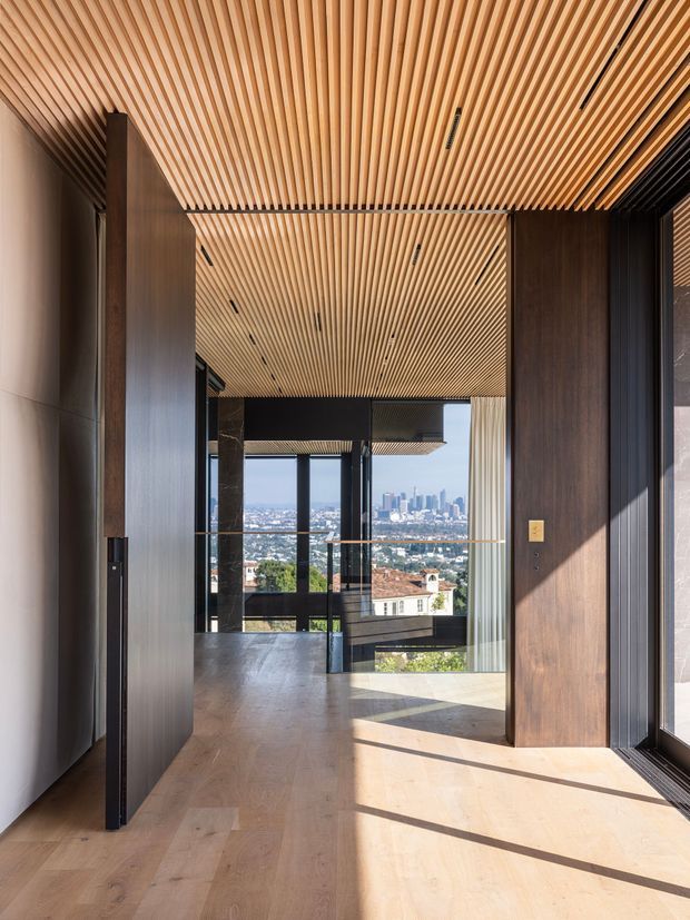 A refined contemporary interior passage or entry zone with a warm timber slatted ceiling, matching timber wall panels and pale timber flooring. Dark vertical joinery and framing contrast with the natural wood, while full-height glazing opens to expansive city views. Minimal recessed linear lighting, a glass balustrade and strong sunlight create a calm, architectural atmosphere.